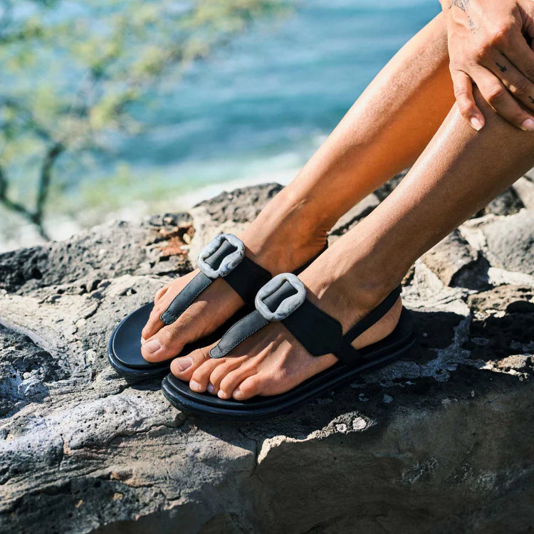 Person wearing black sandals with gray straps on a rocky surface near water.