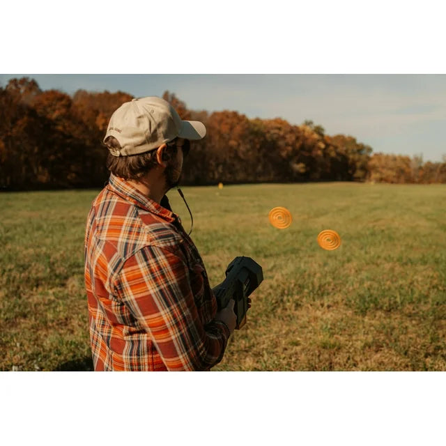 Person in a plaid shirt and cap standing in a field with two orange frisbees.
